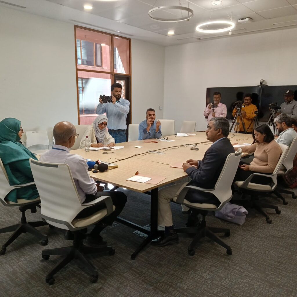 Healthcare professionals discussing Nipah virus risk and public health preparedness during a medical briefing, with laboratory equipment and virus research visuals in a clinical conference setting.