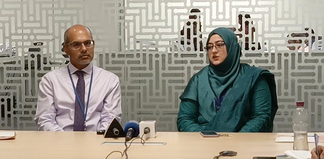Healthcare professionals discussing Nipah virus risk and public health preparedness during a medical briefing, with laboratory equipment and virus research visuals in a clinical conference setting.